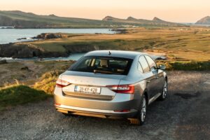 Silver Skoda Superb parked above the coastline near Dunquin with panoramic views of sea cliffs, green fields, and distant peaks.