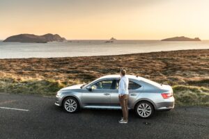 Man standing beside a silver car on Slea Head Drive, gazing at the Blasket Islands silhouetted against a golden Atlantic sunset.