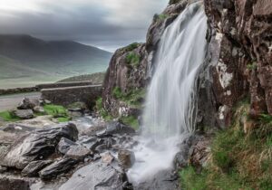 Dramatic waterfall flowing beside a stone bridge along Conor Pass, surrounded by rocky cliffs and lush green hills in County Kerry, Ireland.