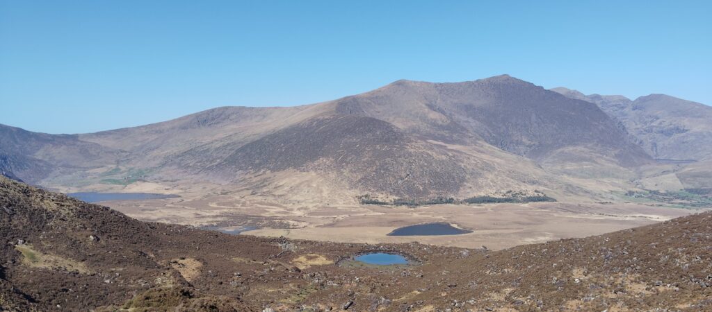 Sweeping vista from Conor Pass showing rugged mountains, glacial lakes, and moorland terrain under a clear blue sky in County Kerry.