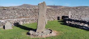 Ornately carved early Christian cross slab at the Riasc monastic site, surrounded by dry-stone walls and rolling hills in County Kerry.