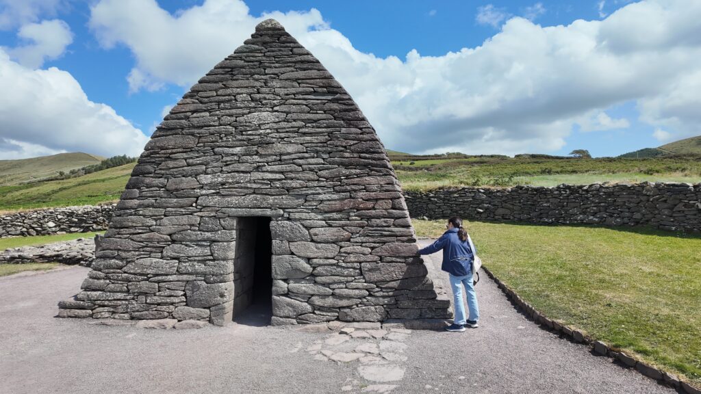 Visitor standing beside the exterior wall of Gallarus Oratory, illustrating the scale of the early medieval stone church on the Dingle Peninsula.