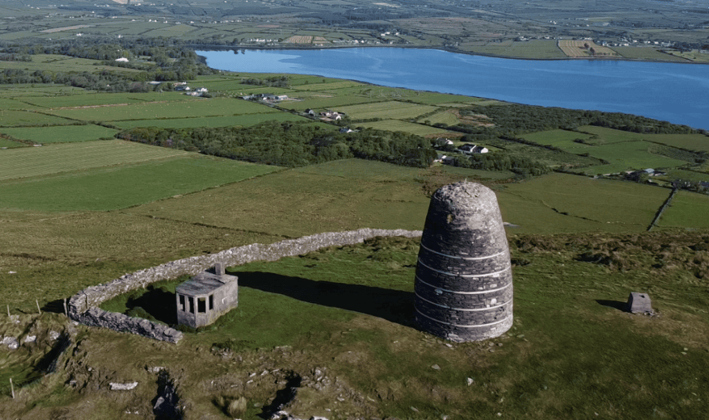 Aerial view of Eask Tower and its surrounding stone enclosure overlooking Dingle Harbour and the patchwork fields of the Dingle Peninsula.