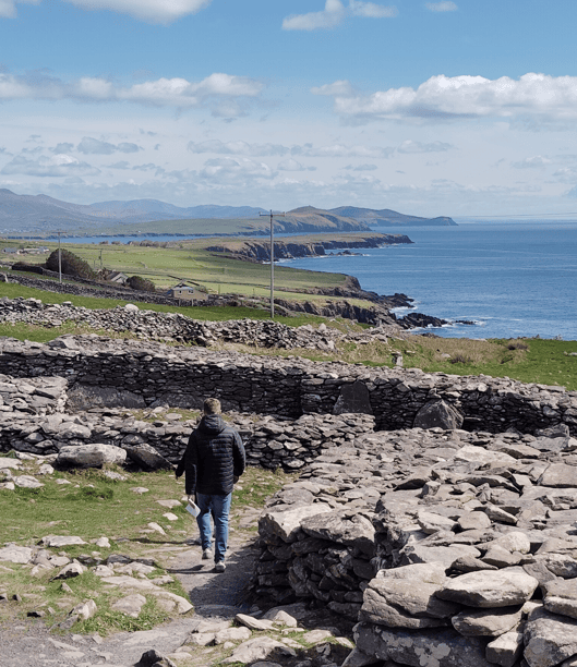 Visitor walking among Fahan stone ruins overlooking green coastal fields and the Atlantic coastline of the Dingle Peninsula.