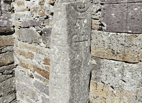 Close view of the Kilmalkedar Ogham Stone showing its weathered surface and carved markings beside the church masonry.