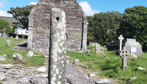 Kilmalkedar Ogham Stone standing in the graveyard with the ruined stone church and carved crosses visible behind it.