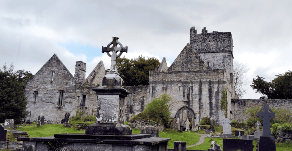 Muckross Abbey ruins with a large Celtic cross in the foreground beneath a cloudy sky in Killarney National Park.
