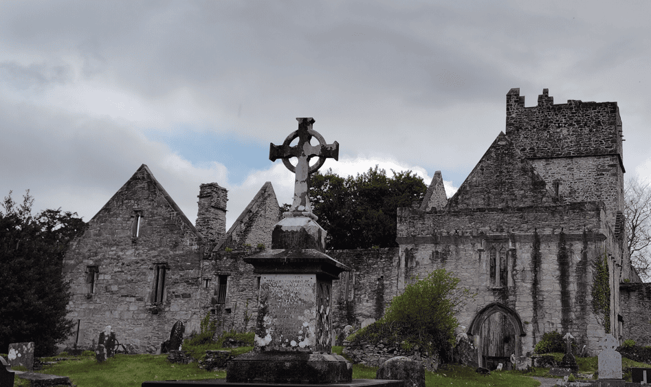 Muckross Abbey ruins with a Celtic cross and stone memorial in the foreground beneath an overcast sky.
