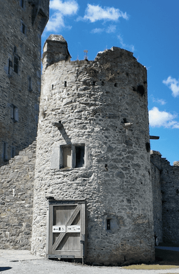 Rounded stone corner tower at Ross Castle showing narrow openings, a doorway and adjoining medieval masonry beneath a blue sky.