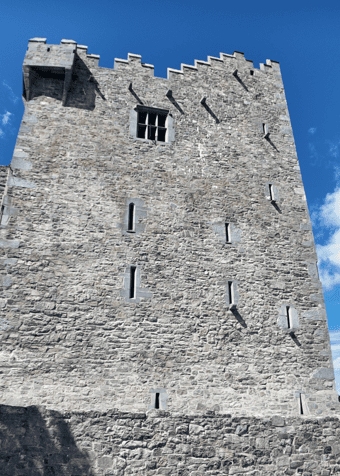 Tall stone tower house of Ross Castle viewed from below, showing narrow openings, battlements and projecting defensive features.