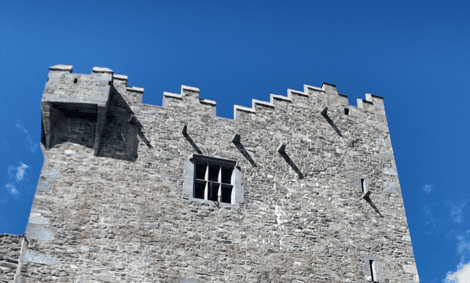 Upper walls of Ross Castle showing the crenellated battlements, projecting corner turret and stone masonry against a blue sky.