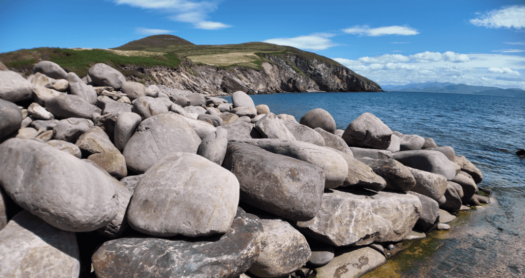 Large rounded boulders along Kilmurry Bay with blue water and steep coastal hills on the southern Dingle Peninsula.