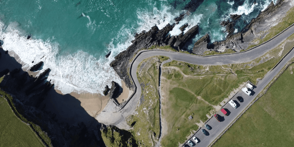 Aerial view of Coumeenoole Beach showing the sandy cove, Atlantic surf, coastal road and parking area beneath steep cliffs.