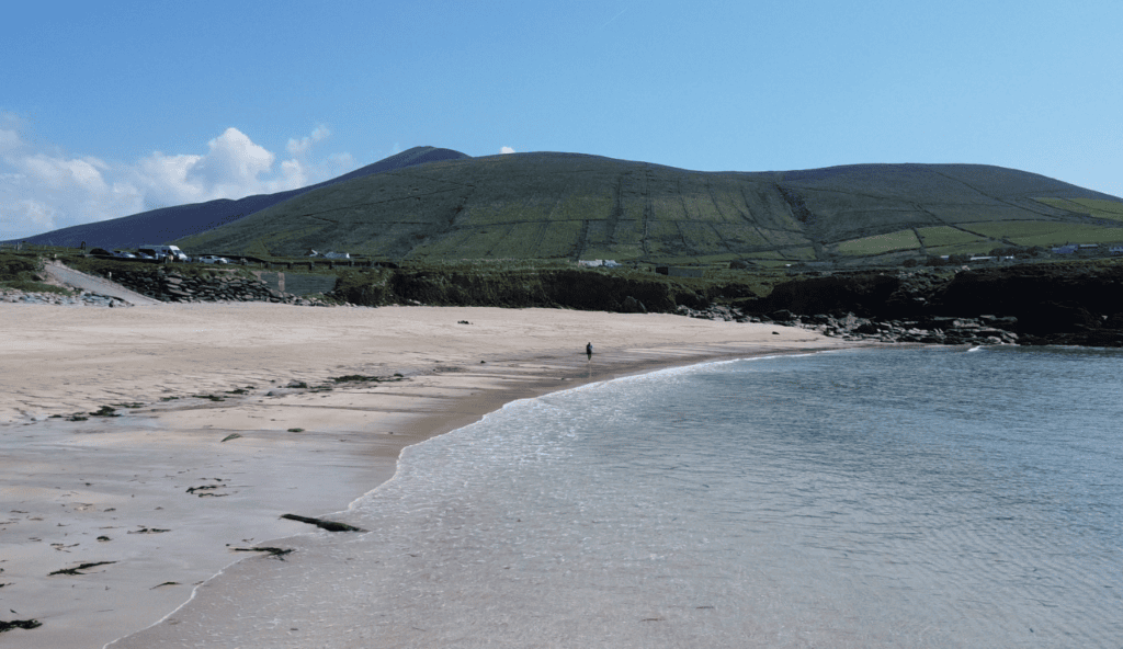 Clogher Strand viewed from the shoreline, with a broad sandy beach, gentle Atlantic waves and green hills rising behind the coast.