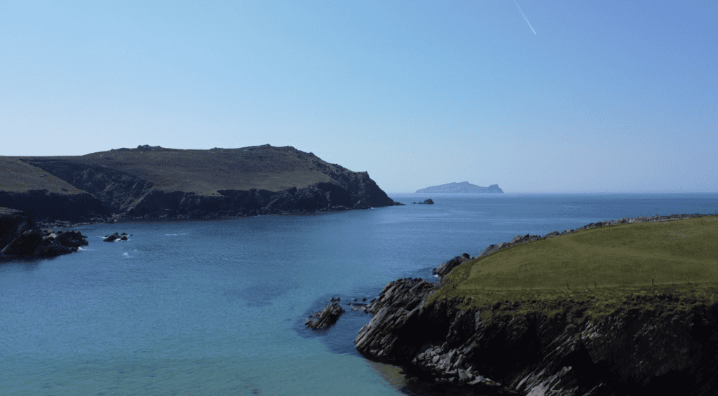 Clogher Strand coastline facing the Atlantic, with rocky headlands, blue ocean and distant islands off the western Dingle Peninsula.