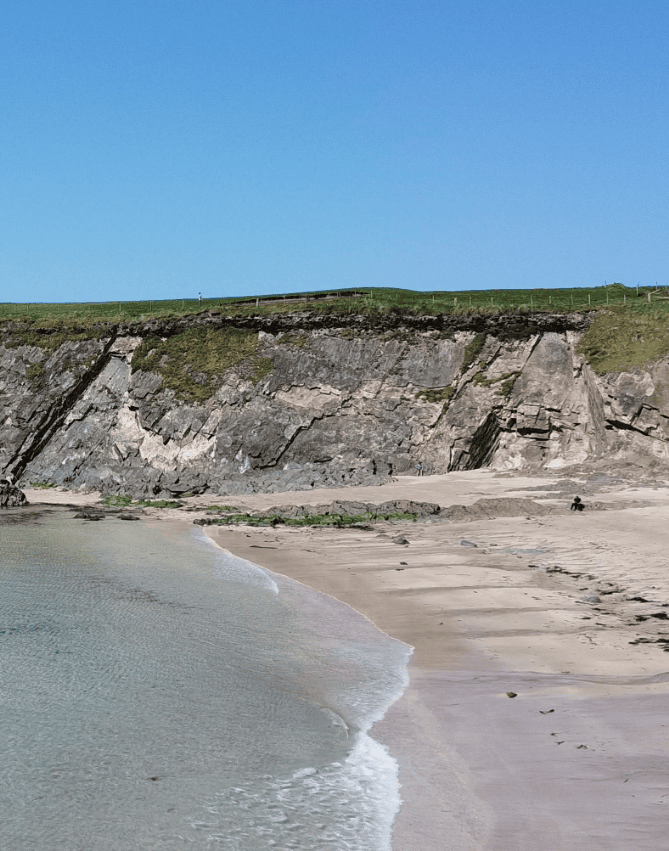 Clogher Strand shoreline beneath exposed rocky cliffs, with a sandy beach and gentle Atlantic waves on the Dingle Peninsula.