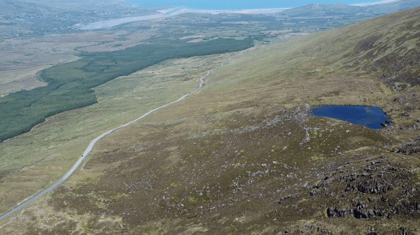 Aerial view across Conor Pass showing the winding mountain road, steep slopes, a small lake and distant coastline.