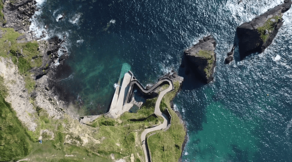 Overhead aerial view of Dunquin Pier showing its winding path, concrete landing area, rocky coastline and clear Atlantic water.