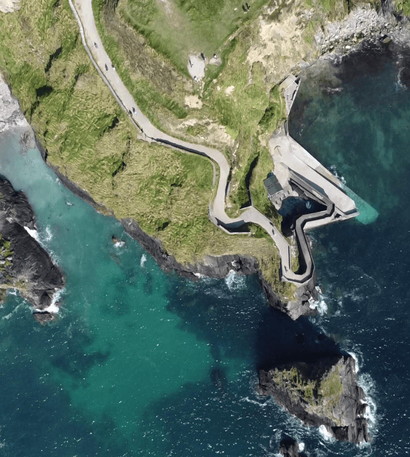 Aerial view of Dunquin Pier showing the steep winding access path descending to the concrete pier beside clear Atlantic waters.