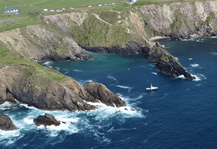 Rugged sea cliffs and rocky Atlantic coastline at Dún Chaoin, with a small boat visible in the sheltered water near Dunquin Pier.