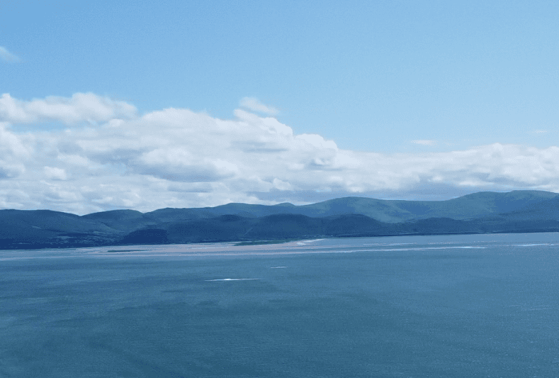 View across Dingle Bay from Rossbeigh Strand, with blue water and the mountains of the Dingle Peninsula beneath scattered clouds.
