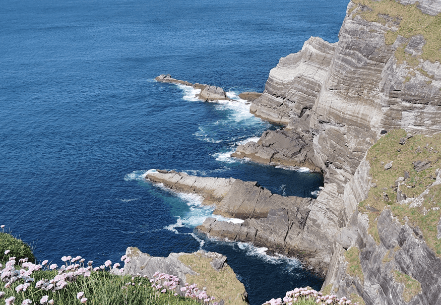 Layered rock ledges at Kerry Cliffs descending into the Atlantic Ocean, with waves breaking against the exposed coastline.