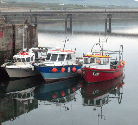Three small fishing boats moored in the calm harbour at Portmagee, County Kerry, with their reflections visible in the water.