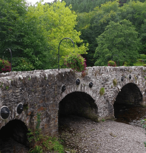 Historic stone Behy Bridge crossing the River Behy near Glenbeigh, County Kerry, with multiple arches and wooded banks.