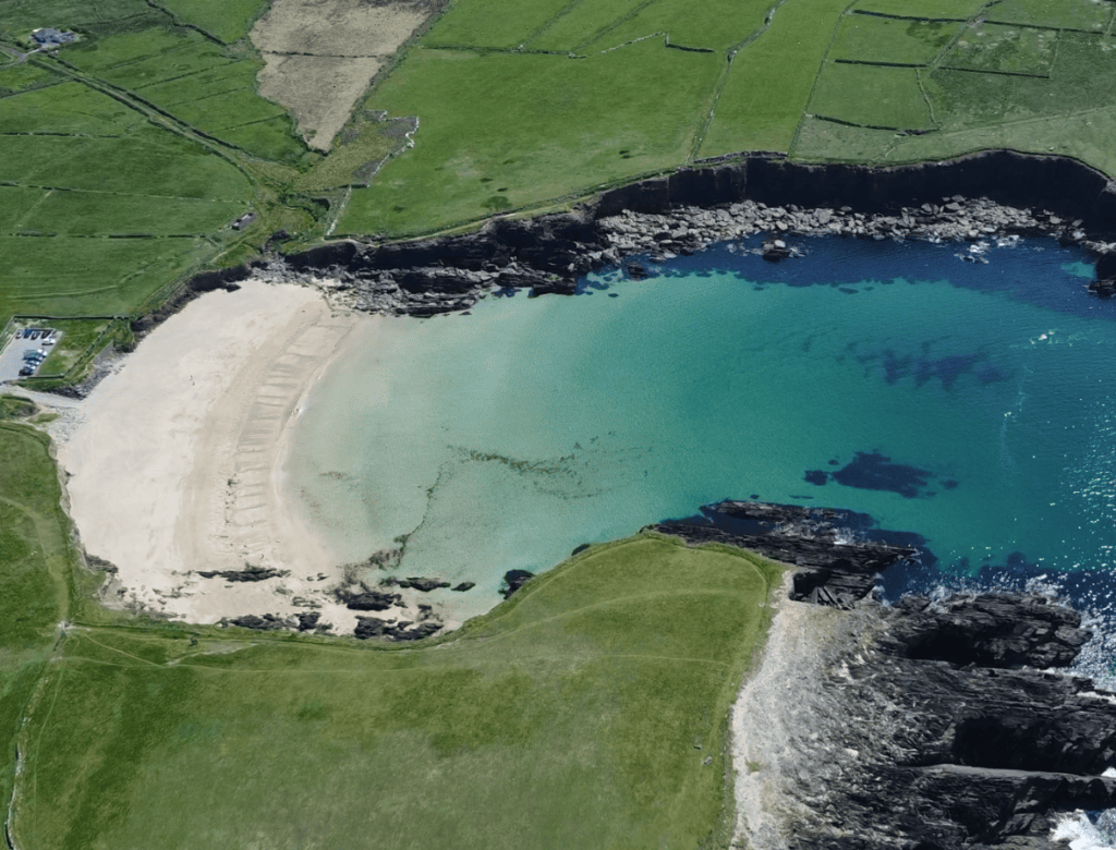 Aerial view of Clogher Strand’s curved sandy beach, turquoise water, rocky shoreline and surrounding green fields.